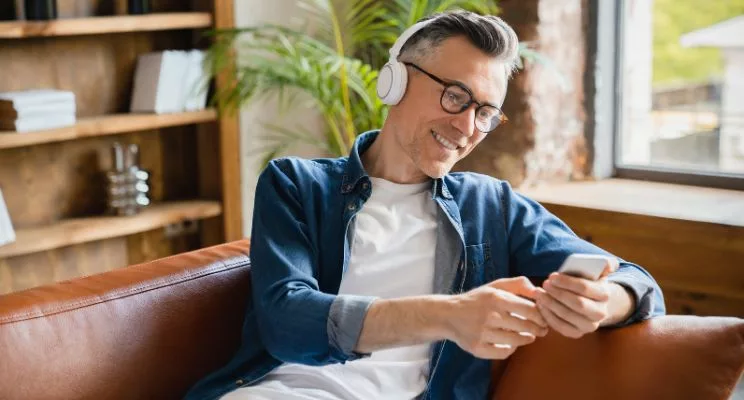 Man with glasses wearing headphones and looking at phone while sitting on couch